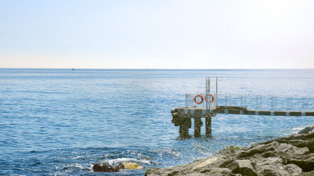 A solitary pier with lifebuoys stretches into the calm, endless blue sea, evoking themes of safety, solitude, and the vastness of nature on a bright, peaceful day.の写真素材