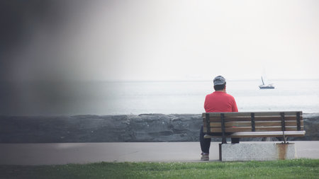 A solitary man sits on a bench, gazing at the calm sea while a sailboat drifts in the distance. The serene scene embodies reflection, peace, and escape from the everyday.の写真素材