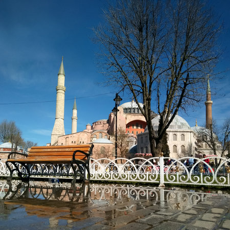 View of Hagia Sophia Mosque with a bench and tree in the foreground, reflecting on wet pavement,...