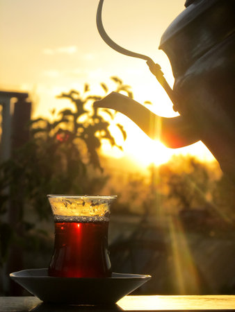 A steaming glass of Turkish tea and a vintage kettle silhouette basking in the warm glow of a golden sunset, evoking a sense of tradition and tranquilityの写真素材