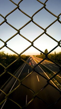 A highway seen through a chain-link fence at sunset, representing the contrast between confidence and freedom, as well as the endless journey toward possibilities.の写真素材
