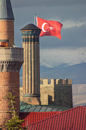 A dramatic view of the Twin Minaret Madrasa, Erzurum Castle, and a Turkish flag waving against a scenic mountainous backdrop, showcasing Ottoman heritageの写真素材