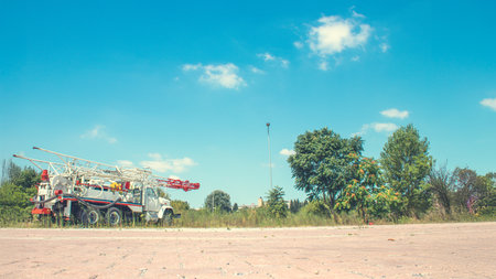 A vintage truck with a crane arm rests on a paved lot surrounded by greenery, blending industrial charm and natural serenity under a clear blue sky with scattered clouds.の写真素材