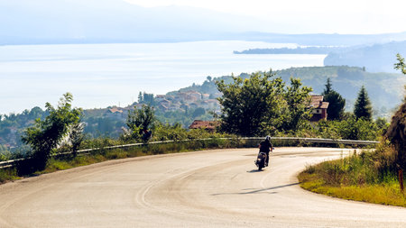 A motorcyclist enjoys freedom on a winding road surrounded by lush greenery, with a tranquil lake and distant mountains in the background, symbolizing travel and exploration.の写真素材