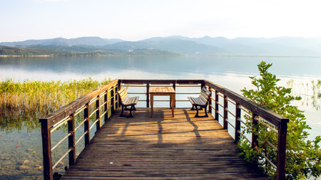 A serene wooden pier with benches and table on Sapanca Lake, reflecting tranquility and a peaceful natural landscape with lush greenery and clear water.の写真素材