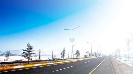 A scenic winter road with frosted trees, distant mountains, and a clear blue sky. A tranquil and crisp atmosphere highlights the serene beauty of winter.の写真素材
