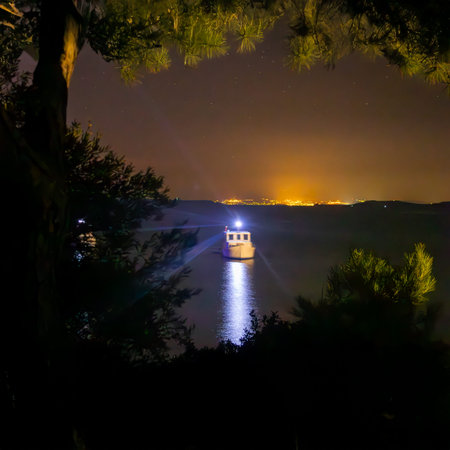 A peaceful night sea scene from Edremit, Balikesir, showcasing a glowing boat on the calm water, surrounded by natural silhouettes and distant city lights.の写真素材