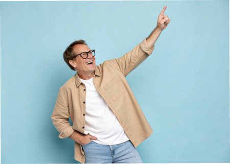 Happy young man in casual clothes and eyeglasses pointing up on blue backgroundの素材
