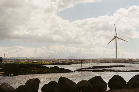 A wide coastal landscape featuring a large wind turbine near the shoreline, calm water, and a...