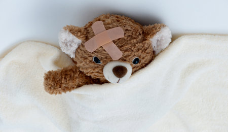 Sick teddy bear toy with patch in on head lying in bedの写真素材