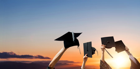 Four hands holding graduation hats against sunrise skyの写真素材