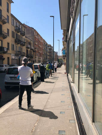 A long line of people, wearing a surgical mask and spacing 1 meter apart, to enter in a supermarket due to coronavirus, in an Italian street in a city in the north of the country ina sunny dayのeditorial素材
