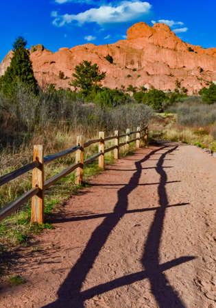 A wooden fence with a shadow from the sun on a background of red mountain formations along which a person walks. Colorado Springs area is Garden of the Gods, National Natural Landmarkの写真素材