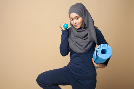 A young beautiful hijab woman is holding  a fitness mat and dumbbell on a yellow background .の写真素材