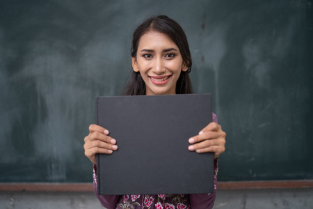 a teacher is holding a blank black book in fornt of a blackboard in a class roomの写真素材