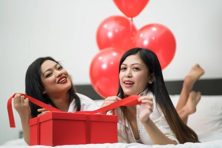 Beautiful young woman and his friend with balloons red and box on bed. Valentine's day celebrationの写真素材
