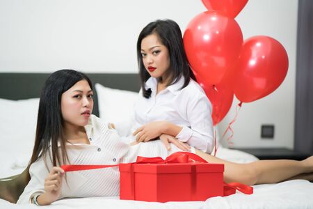 Beautiful young woman and his friend with balloons red and box on bed. Valentine's day celebrationの写真素材