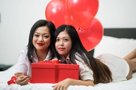 Beautiful young woman and his friend with balloons red and box on bed. Valentine's day celebrationの写真素材