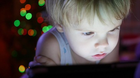 The child looks to the tablet lying on bed.On the background lights garlandsの写真素材