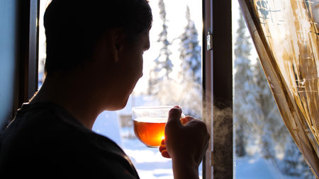 Winter frosty day. The guy early in the morning standing at the window and drinking hot tea.