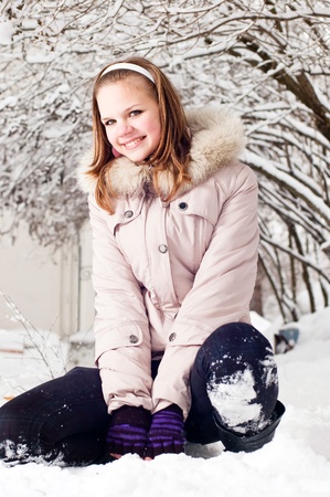 Beautiful young girl sits on to snow in warm clothes in winter outdoorsの写真素材