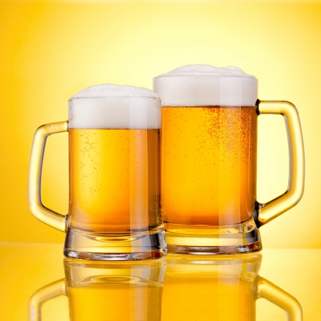 Two Mugs of fresh beer with cap of foam, isolated on yellow backgroundの写真素材