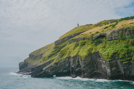 White Lighthouse on a Hill and a Steep Coastal Cliff of Jeju Island, Korea, Rising Above the Blue Wavesの写真素材