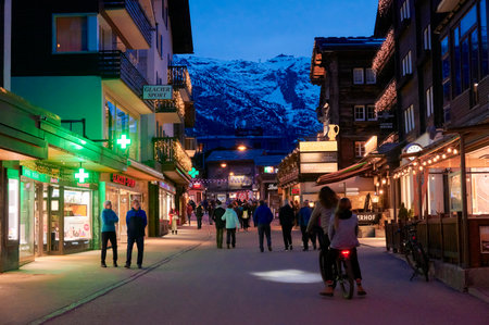 ZERMATT, SWITZERLAND - APR 11, 2022: Tourists walking and sightseeing on a street in the city of Zermatt at sunset.のeditorial素材