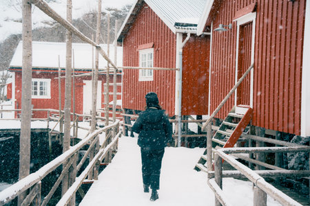 Person walking on a snowy path between red wooden cabins in winter.Nusfjord, Lofoten, Norway.の写真素材