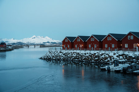 Red wooden rorbuer cabins in snowy Leknes, Lofoten, Norway, glow in golden sunrise light near a...