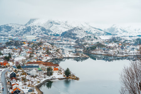 Picturesque harbor view of Henningsvaer, Lofoten, Norway, with fishing boats, traditional buildings, and dramatic snow-covered mountains reflected in water.の写真素材