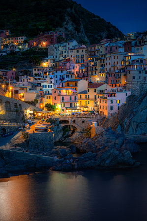 Nighttime view of Manarolaâs pastel cliffside buildings illuminated by warm lights, reflecting off the Ligurian Sea in Cinque Terre.の写真素材