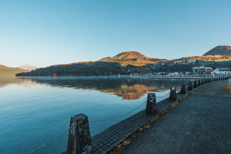 Peaceful morning along the waterfront promenade in Moto-Hakone, overlooking Lake Ashi and sunlit mountains with village buildings.の写真素材