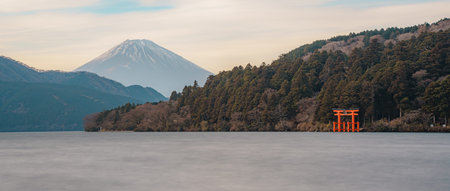 Scenic view of a traditional red torii gate standing in Lake Ashi with forested hills in the...