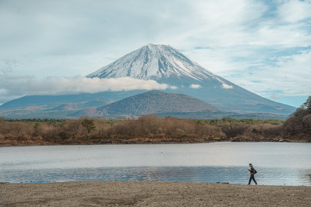 Fujikawaguchiko, Japan - Dec 12, 2023: Majestic Mount Fuji covered in snow looms over tranquil Lake Shoji, reflecting clouds and blue sky, framed by forested shores.の写真素材
