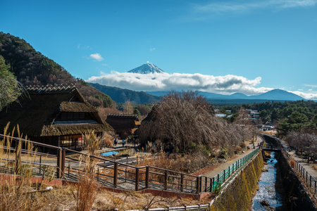 Charming traditional village with thatched-roof houses set against Mount Fuji's snowy peak, surrounded by forests and blue skies in Saiko Iyashi no Sato Nenba.の写真素材