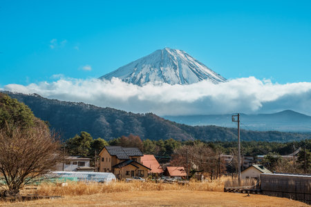 Charming traditional village with thatched-roof houses set against Mount Fuji's snowy peak, surrounded by forests and blue skies.の写真素材
