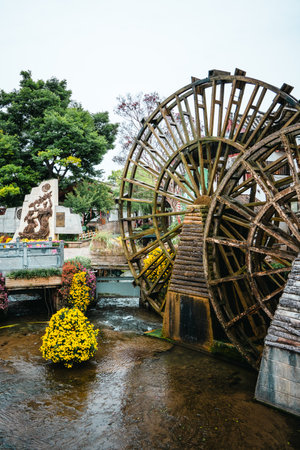 A large rustic waterwheel turns slowly in a stream flanked by yellow flowers and heritage sculptures...