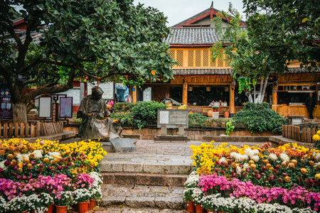 A bronze statue sits beneath a tree surrounded by vibrant potted chrysanthemums in a quiet courtyard of Lijiang Old Town, Yunnan.の写真素材