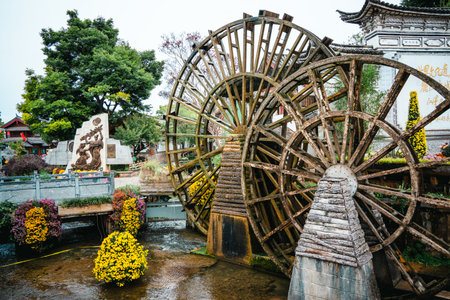 A large rustic waterwheel turns slowly in a stream flanked by yellow flowers and heritage sculptures in Lijiang Old Town.の写真素材