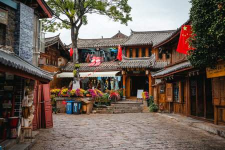A quiet square in Lijiang Old Town features wooden shops, potted flowers, and traditional Chinese architecture with flags overhead.の写真素材