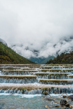 Scenic terraced waterfalls flowing into turquoise pools at Blue Moon Valley, surrounded by lush green hills and misty skies.の写真素材