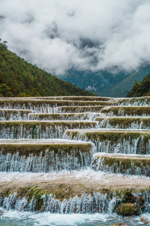 Scenic terraced waterfalls flowing into turquoise pools at Blue Moon Valley, surrounded by lush green hills and misty skies.の写真素材