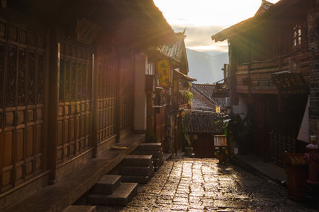Sunlight filters through a narrow alleyway lined with wooden buildings and cobblestone in Lijiang Old Town, Yunnan, evoking historic charm.の写真素材