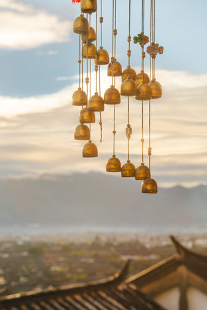 ells and prayer plaques hang from a wooden structure overlooking rooftops and mountains at sunrise in Lijiang Old Town, Yunnan.の写真素材