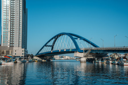 Fishing boats docked near tall skyscrapers and a blue arch bridge at Sokcho harbor under clear blue sky.の写真素材
