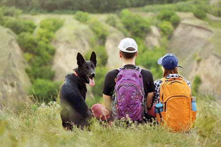 Back view of a sitting at the hill hiking couple with a dogの写真素材