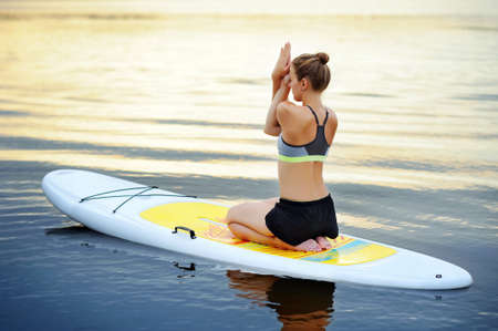 Woman practicing sitting variation of eagle yoga pose on paddle board