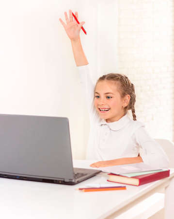 Smiling schoolgirl sitting at a table with a laptop and pulls her hand up
