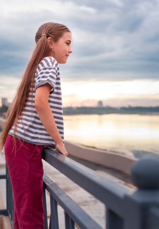 Adorable little girl standing on the bridge over the river at sunsetの写真素材